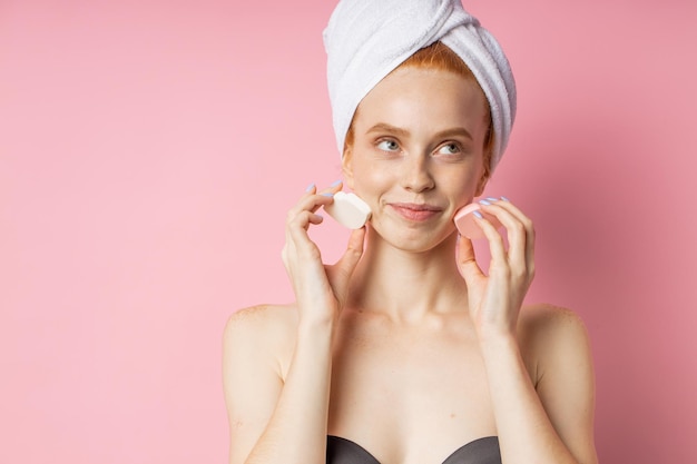 Gorgeous young caucasian woman with towel on head holding two cosmetic sponges, with pleasure, smiles widely, happy with her clean skin, posing against pink studio wall.