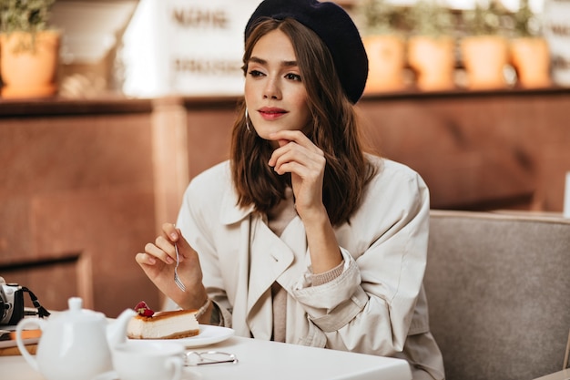 Gorgeous young Parisienne with dark wavy hairstyle, red lips, beret, beige trench coat, sitting at city cafe terrace, having cheesecake and tea, looking away