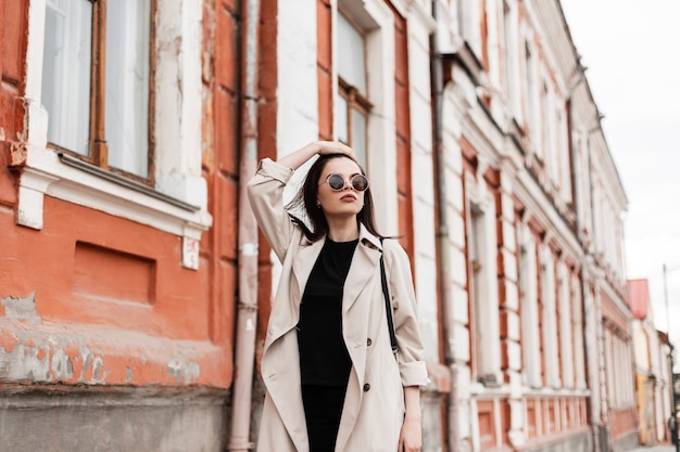 Gorgeous young woman in fashion black clothes in stylish sunglasses in beige trench coat walks and straightens hair. Attractive girl model in casual outfit walk on city street near vintage building.