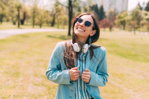 A gorgeous young woman is smiling at the camera with her sunglasses on and headphones in park