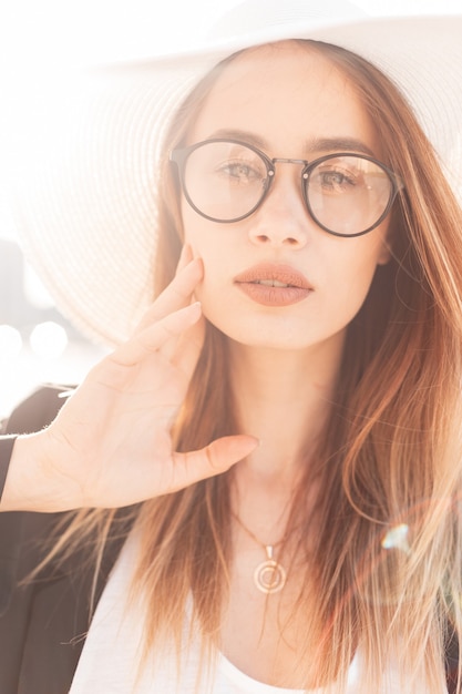 Gorgeous young woman with beautiful lips in fashionable glasses in straw white elegant stylish hat in black trendy jacket posing in bright sunlight. Charming pretty girl at sunset. Fashion portrait.