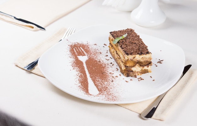 Gourmet Slice of Tasty Chocolate Cake on White Plate with Fork Outline Using Cocoa Powder on the Side. Served on White Dining Table.