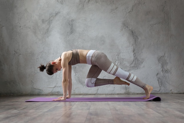 Graceful young brunette woman doing stretching and yoga poses at home or yoga studio Attractive Girl wearing grey pants and top working out indoor