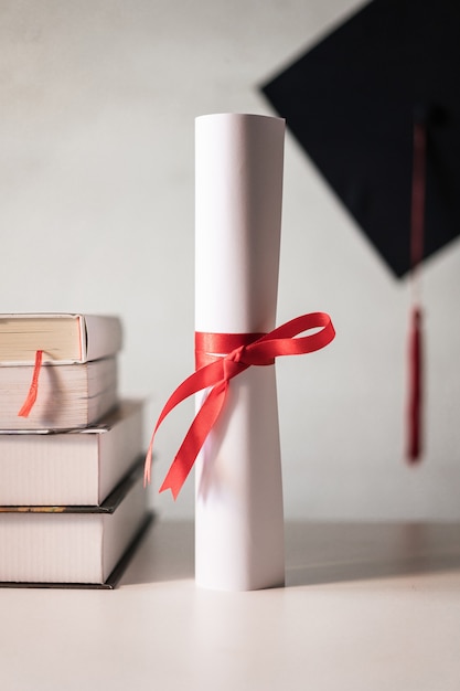 A graduation hat or mortarboard and diploma certificate paper tied with red ribbon on a stack of  books with empty space slightly undersaturated with vignette for vintage effect