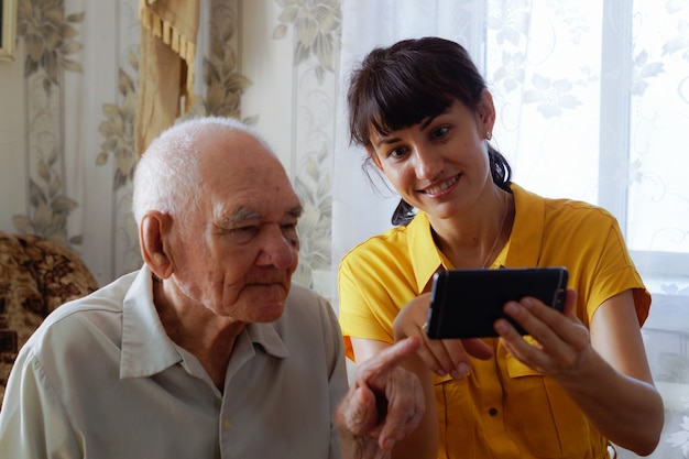 A grandfather and an adult granddaughter are sitting on the sofa and looking at their mobile phone