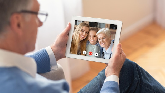 Grandfather Having Video Call With Family Sitting With Digital Tablet Talking With Senior Wife, Adult Daughter And Little Grandchild At Home. Distance Family Communication Concept. Panorama, Cropped