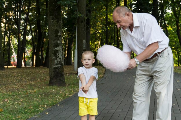 Grandfather lovingly and carefully treats and feeds his little grandson with pink cotton candy in an amusement park A pensioner and a little boy spend their free time on weekends