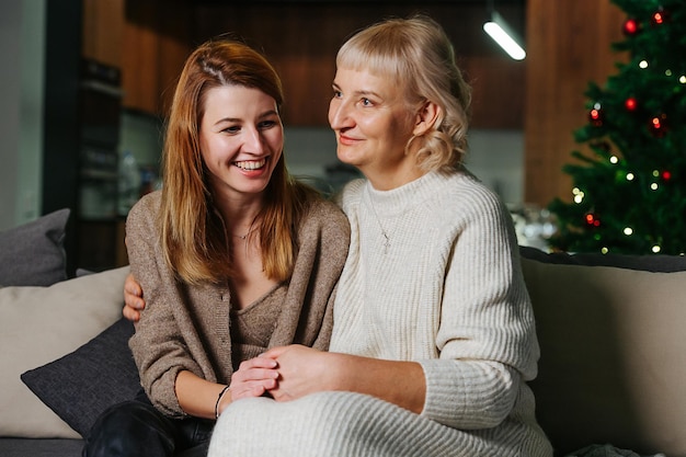 Grandma and her adult daughter sitting on a couch, holding hands. Christmas tree in background.