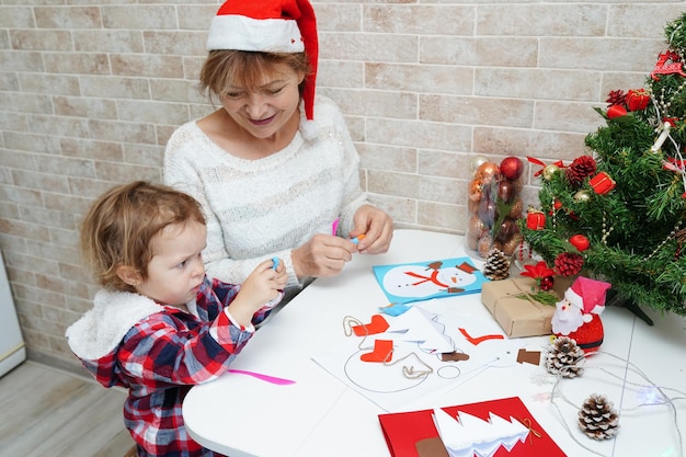 Grandmother and child girl in Santa hat making christmas greeting card with snowman