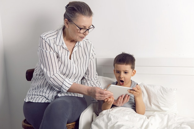 Grandmother in glasses and a white shirt reads a tablet to her grandson lying on the bed in a white children's room