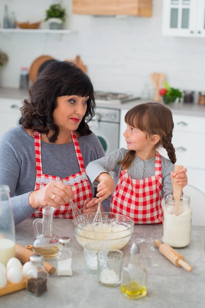 Grandmother and granddaughter in checkered aprons are cooking in the kitchen and looking at each other.