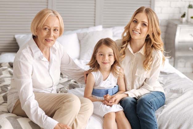 Grandmother, mother and her little daughter sitting on the bed and bonding to each other, while both women hugging a child and smiling.