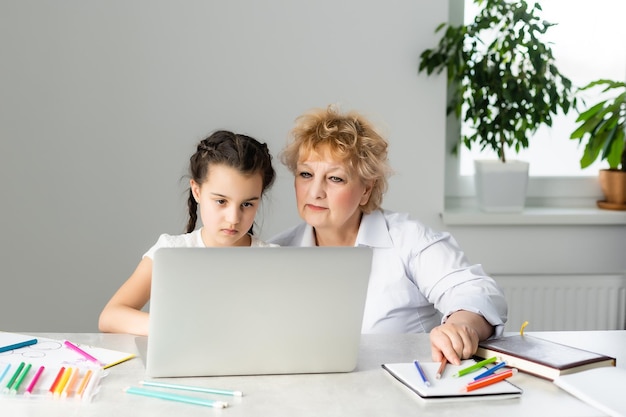 Grandmother teaching to granddaughter with the help of the computer. Worried old teacher helping girl studying and doing homework on laptop at home, private lesson.