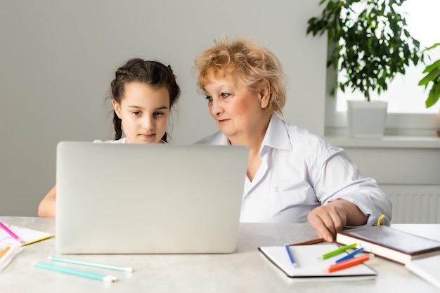 Grandmother teaching to granddaughter with the help of the computer. Worried old teacher helping girl studying and doing homework on laptop at home, private lesson.