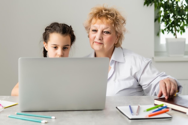 Grandmother teaching to granddaughter with the help of the computer. Worried old teacher helping girl studying and doing homework on laptop at home, private lesson.