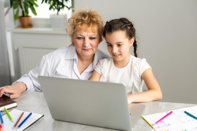 Grandmother teaching to granddaughter with the help of the computer. Worried old teacher helping girl studying and doing homework on laptop at home, private lesson.