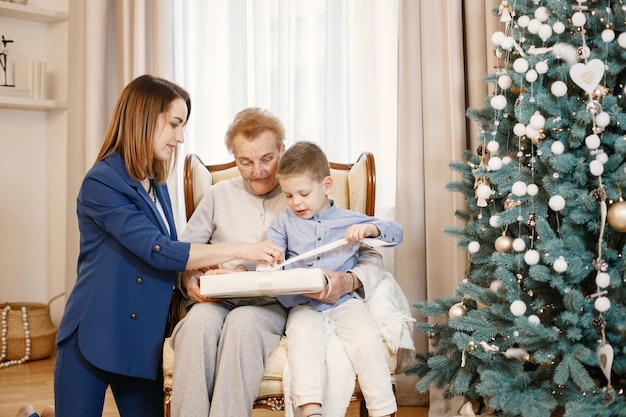 Grandmother with her daughter and grandson on Christmas day. Women and little boy opening a gift. Women and boy wearing beige and blue clothes.