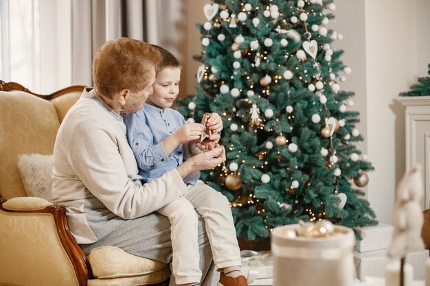 Grandmother with her grandson on Christmas day. Old woman and little boy decorating Christmas tree. Woman and boy wearing beige and blue clothes.