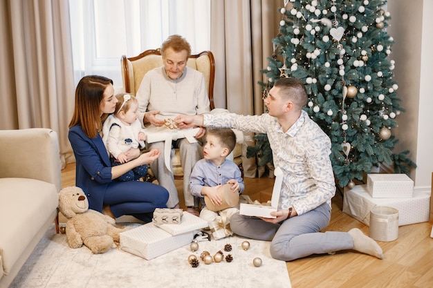 Grandmother with her grandson, granddaughter, daughter and son on Christmas day. Old woman sitting on a chair and other is going to decorate Christmas tree. Family wearing beige and blue clothes.