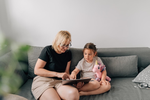 Granny and preschool girl using tablet together at home on the sofa family togetherness time