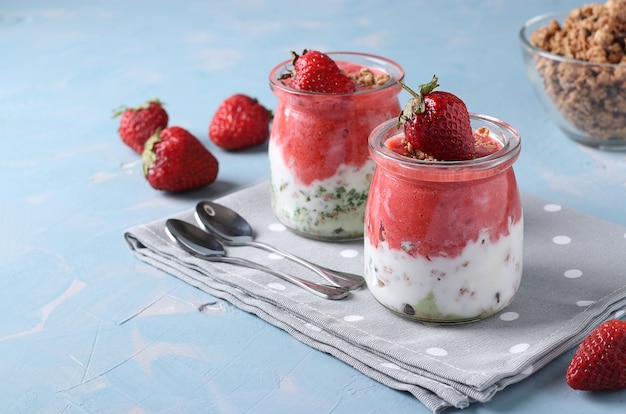 Granola crispy with strawberry, spinach, and natural yogurt, a delicious and healthy breakfast in glass jars on blue background. Horizontal format