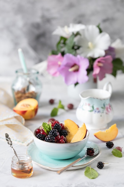 Granola or muesli with berries and fruits for a healthy breakfast in the morning with a bouquet of summer flowers on a light table.