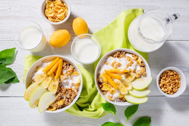 Granola and vegetarian yogurt with slices of apple, apricot, banana and a jug of milk on a white wooden surface. Healthy breakfast concept. Top view