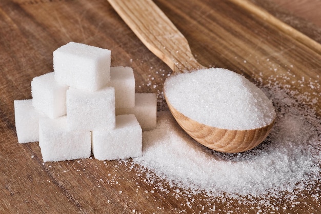 Granulated sugar in wooden spoon and sugar cubes stacked in pyramid on wooden background