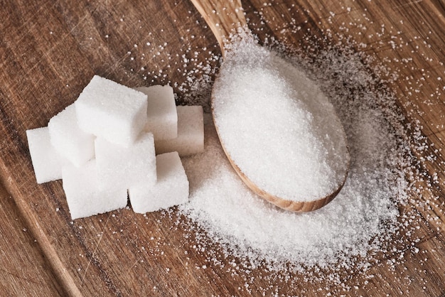 Granulated sugar in wooden spoon and sugar cubes stacked in pyramid on wooden background