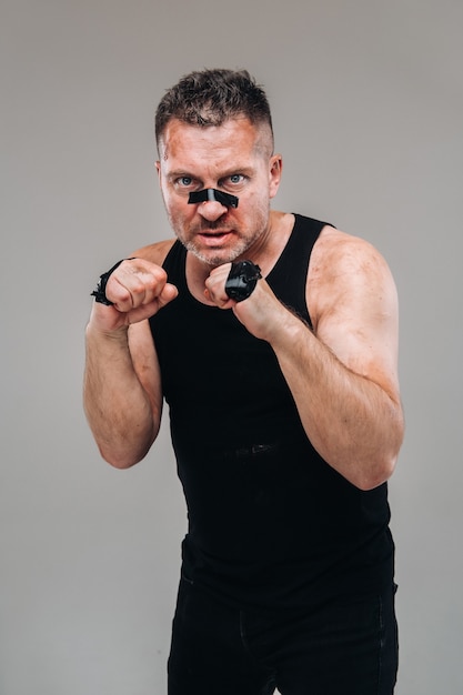 On a gray background stands a battered man in a black T shirt looking like a fighter and preparing for a fight.