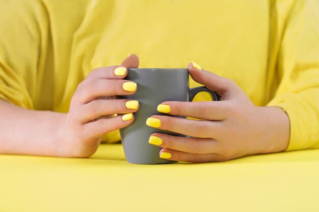 Photo gray cup in hands with yellow manicure on yellow table. colors of the year 2021. illuminating and ultimate gray.