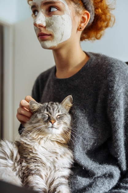 Gray fluffy tabby cat sits on arms teenage girl wearing cosmetic mask