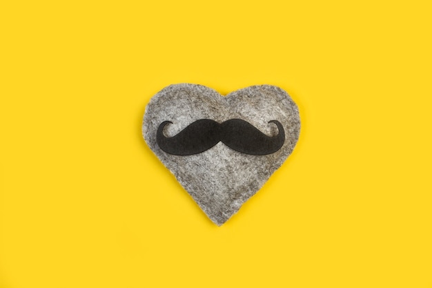 A gray textile heart with a black mustache on a yellow background