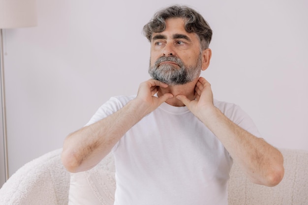 Photo grayhaired man has a severe sore throat senior experienced male sitting on sofa holding his hand by the throat inflammation and sore throat