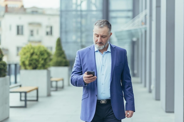 Grayhaired senior handsome man businessman in suit walking near office center on street holding