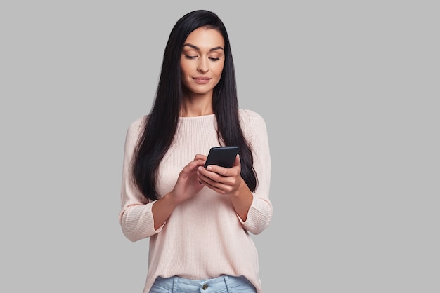 Great app! Attractive young woman smiling and using her smart phone while standing against grey background