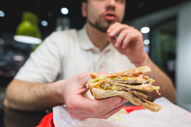 Great appetizing sandwich in the hands of a young man. The man feeds fast food at the restaurant.