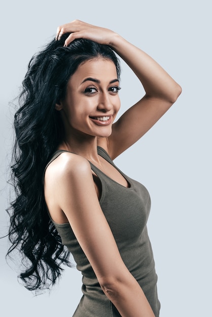 Great hairstyle! Attractive young woman in casual wear holding her hand in hair and looking at camera while standing against grey background