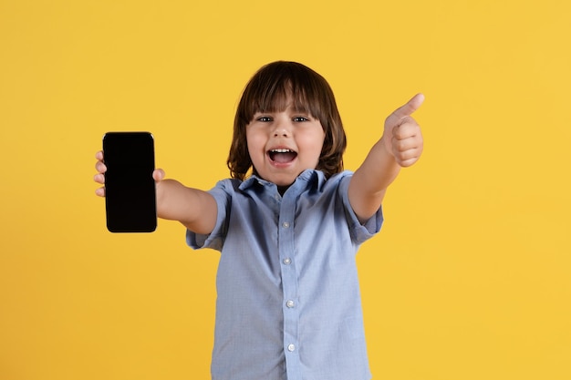 Great kids content Excited little boy showing smartphone with blank screen to camera and gesturing thumb up