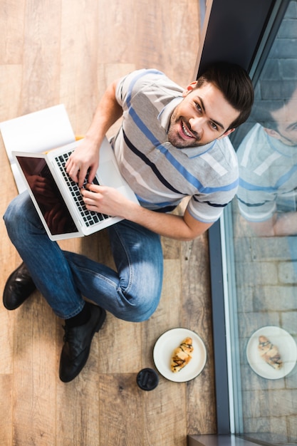 Great mood. Top view of a delighted positive nice man sitting on the floor and looking at you while holding his laptop