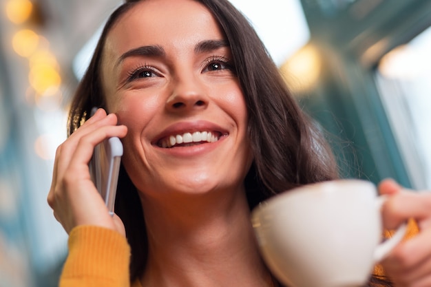 Great news. Close up of attractive glad brunette woman holding phone while smiling and drinking tea