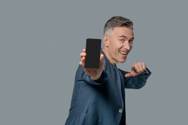 Great smartphone. Happy otimistin man in business jacket showing smartphone screen and pointing with finger on gray background