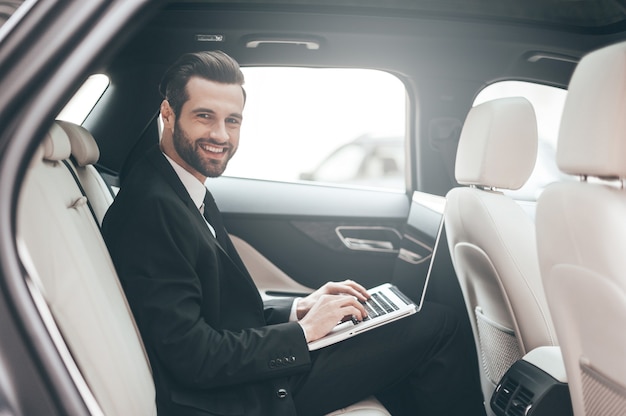 Great solutions every day. Confident young businessman working on his laptop and looking at camera while sitting in the car