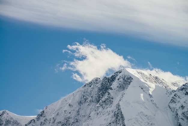 Great view to high snowy mountain peaked top with low cloud under cirrus clouds in sky. Low clouds on big snow covered mountains with sharp pinnacle in sunshine. White-snow pointy peak in sunlight.