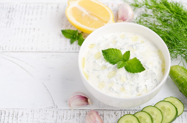 Greek dip sauce or dressing tzatziki and ingredients decorated with olive oil and mint on white wooden table