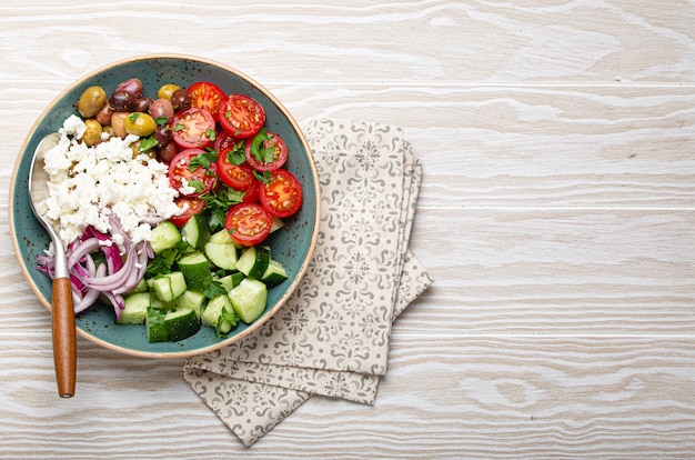 Greek mediterranean salad with tomatoes, feta cheese, cucumber, whole olives and red onion in blue ceramic plate on white wooden background from above, traditional appetizer of Greece with copy space