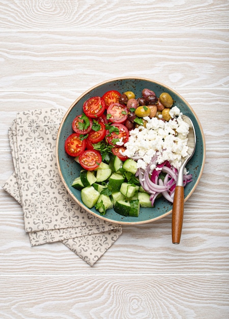 Greek mediterranean salad with tomatoes, feta cheese, cucumber, whole olives and red onion in blue ceramic plate on white wooden background from above, traditional appetizer of Greece