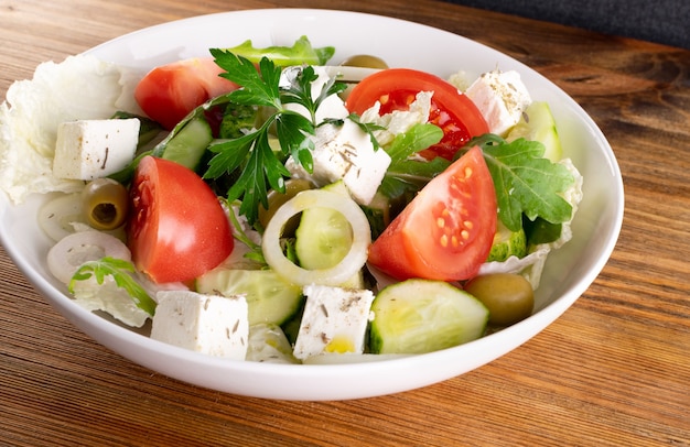 Greek salad or horiatiki with large pieces of tomatoes, cucumbers, onion, feta cheese and olives in white bowl closeup. Village salad with diced mozzarella, arugula, parsley, spices and olive oil