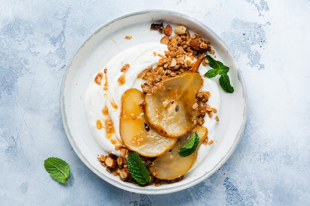 Greek yogurt with caramelized pear, granola, nuts and melted sugar for a wholesome breakfast on a gray ceramic plate. Rustic style. Top view.