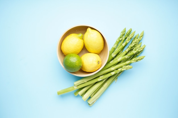 Green asparagus and lemons in a bowl on a blue surface, flat lay.